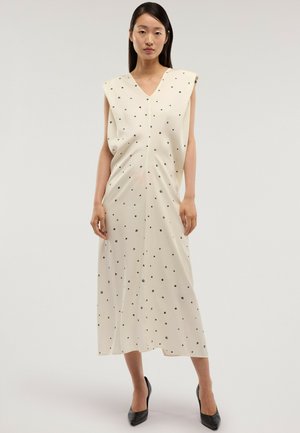 Kvinde iført en ærmeløs cremefarvet midi-kjole med sorte polkaprikker og sorte spidse hæle, stående mod en ensfarvet baggrund.