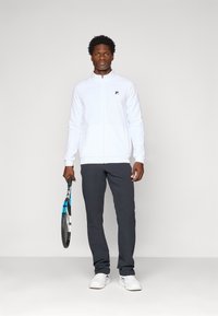 Jachetă albă cu fermoar, dintr-un material texturat, logo negru, asociată cu pantaloni gri închis și adidași albi, ținând o rachetă de tenis.