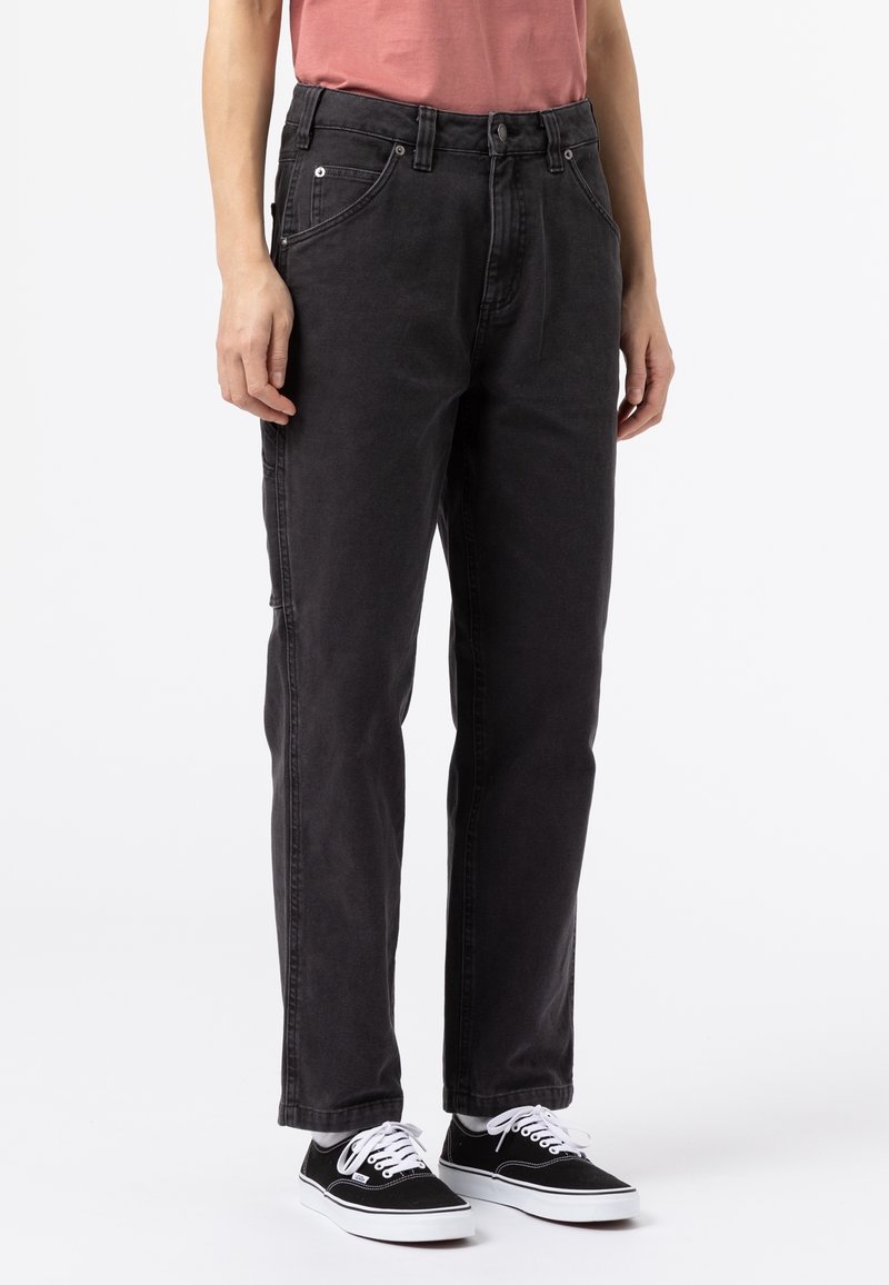 Personne portant un jean droit noir délavé, une chemise rouge atténué et des baskets noires avec des lacets blancs sur fond blanc.