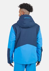 Blauer Skijacke mit Kapuze, die einen navyblauen oberen Abschnitt, strukturierten Stoff und einen kontrastierenden hellblauen unteren Teil aufweist. Das "ROSSIGNOL"-Logo ist sichtbar.