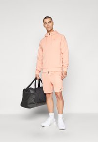 Lys rosa hettegenser og shorts med dragsnor, med et lite svart logo. Hvite sneakers og en svart duffelbag kompletterer antrekket.