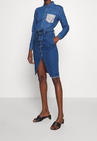 Denim dress with long sleeves, collar, and a sequined pocket. Features a button-down front, belted waist, and a knee-length slit.
