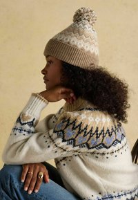 Beżowa dzianinowa czapka z pomponem, z wzorem w zygzak. Białe i jasnoniebieskie akcenty na kremowym swetrze z wzorzystą częścią przy dekolcie.