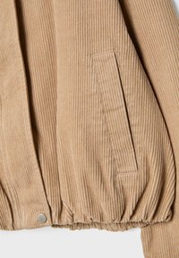 Section d'une veste en velours côtelé beige montrant une texture côtelée verticale, une poche latérale oblique avec rabat, et un ourlet élastique avec bouton-pression métallique.