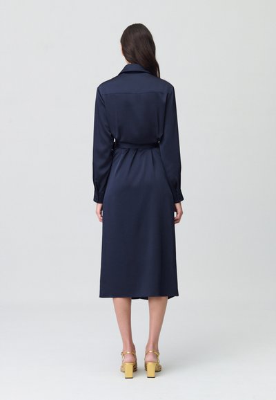 Vestido de satén azul marino con cuello camisero, mangas largas y cinturón en la cintura, con un largo medio y textura suave. Sandalias amarillas de tacón bloque.