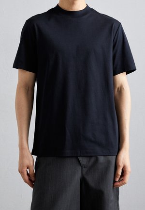 Persona che indossa una maglietta nera semplice a maniche corte con scollo rotondo e pantaloni scuri a righe sottili su sfondo grigio neutro.