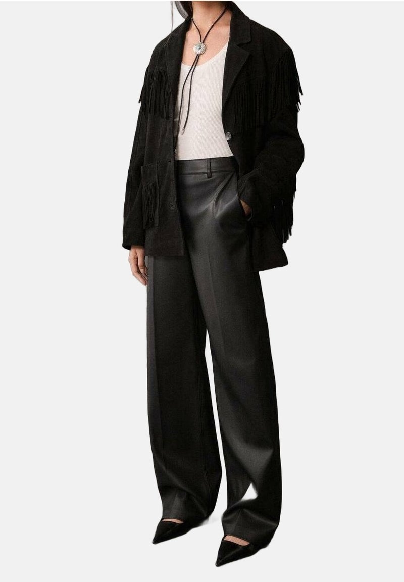 Femme portant une veste noire à franges, un débardeur blanc, un pantalon en cuir noir à jambes larges, des chaussures noires pointues et un collier bolo.