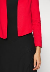 Kvinna som bär en klar röd jacka över en svart klänning med en arm avslappnad vid sin sida mot en enkel vit bakgrund.