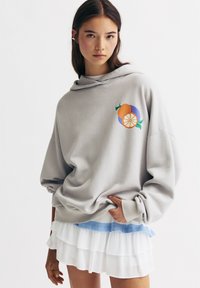 Sweatshirt gris avec capuche, présentant un graphique de fruits coloré sur la poitrine. Jupe blanche superposée avec un bord bleu en dessous.