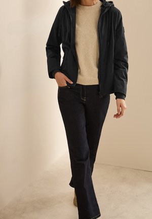 Person trägt eine schwarze Kapuzenjacke mit Reißverschluss, beigen Pullover, schwarze Hose und beigen Schuhe und steht vor einem hellen Hintergrund.