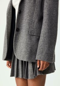 Blazer grigio in lana con due bottoni, taschino sul petto, abbinato a una gonna grigia plissettata. Tessuto testurizzato, taglio moderno e linee pulite.