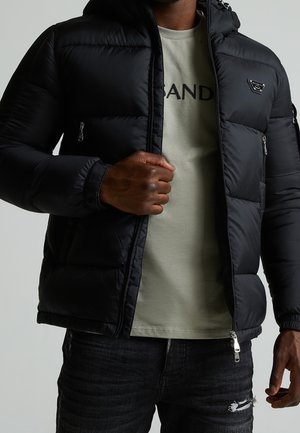 Homme ajustant une veste matelassée noire par-dessus un T-shirt vert clair avec un texte, portant un jean noir déchiré sur un fond neutre.