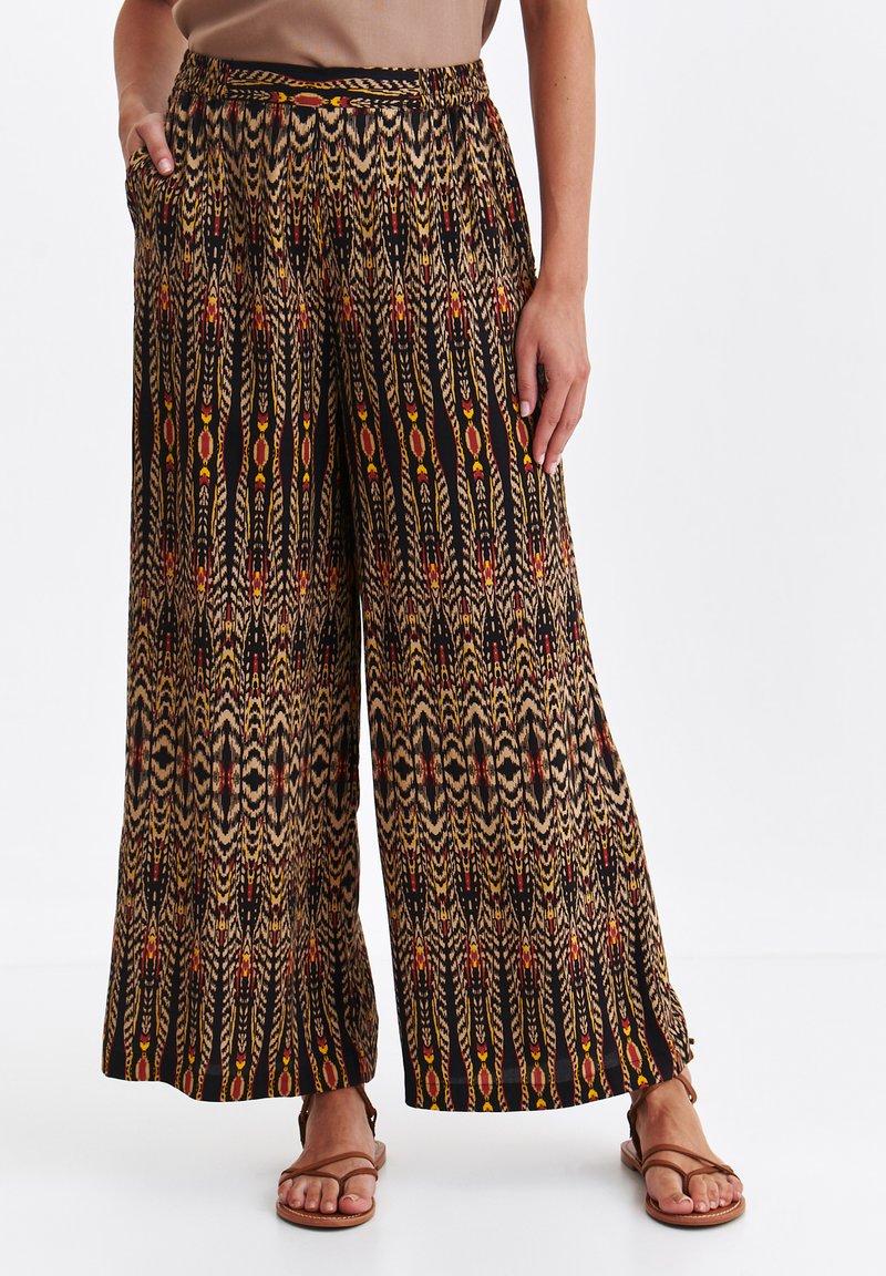 Spodnie o szerokich nogawkach z wielokolorowym, geometrycznym i plemiennym wzorem, z elastycznym pasem, noszone z brązowymi sandałami na paski na białym tle.