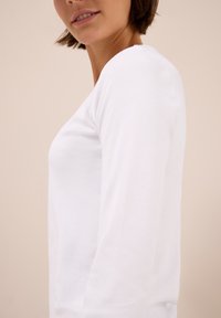 Mujer con una camiseta blanca de manga larga sencilla, contra un fondo beige, mostrada de cuello a la mitad del torso de perfil.