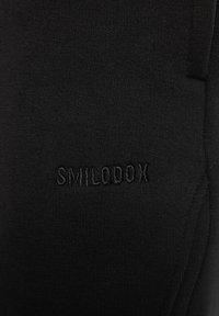 Sorte sportlige leggings lavet af strækbart stof, med et præget "SMILODOX" logo i samme farve på siden.
