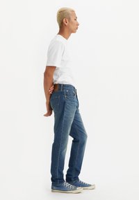 Jeans de mezclilla de lavado claro con corte recto y parche de cuero marrón en la parte posterior. Combinados con una camiseta blanca ajustada y zapatillas deportivas azules.