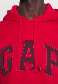 Sudadera roja con el logo bordado de "GAP" en color burdeos oscuro, que presenta un cuello con cordón y una textura de tela. Primer plano de los detalles de la tela y el diseño.
