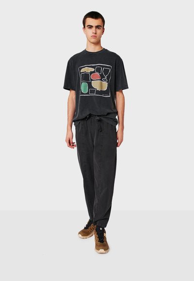 T-shirt en coton noir avec un design graphique abstrait multicolore. Associé à un pantalon noir et des baskets marron avec une semelle texturée.