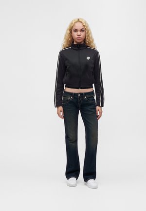 Jeune femme aux cheveux blonds bouclés portant une veste noire zippée, un jean foncé et des baskets blanches, debout devant un fond uni.
