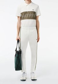 Bílá polokošile s olivově zeleným pruhem a nápisem "LACOSTE", spárovaná s bílými tepláky se zelenými bočními pruhy. Drží zelenou tašku.