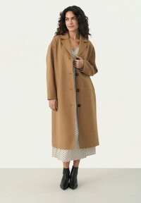 Brauner Wollmischungs-Mantel mit langen Ärmeln, Knopfverschluss und seitlichen Taschen, über einem knielangen gepunkteten Kleid und schwarzen Ankle Boots getragen.