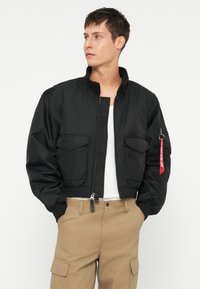Giacca bomber nera con chiusura a zip, polsini elasticizzati e due tasche frontali. Presenta un'etichetta di stoffa rossa sulla manica sinistra. Materiale liscio e leggero.