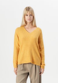 Maglione a V, con maniche lunghe in tessuto a costine giallo, dalla vestibilità comoda e spalle scese, abbinato a pantaloni beigi larghi.