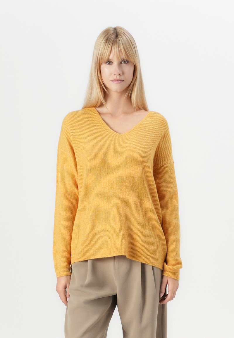 Maglione a V, con maniche lunghe in tessuto a costine giallo, dalla vestibilità comoda e spalle scese, abbinato a pantaloni beigi larghi.