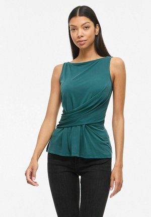 Femme aux longs cheveux noirs portant un haut sans manches bleu sarcelle avec un détail drapé à la taille et un pantalon noir, debout devant un fond blanc.