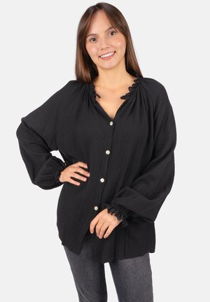 Femme souriante, portant un chemisier noir ample à boutons avec des poignets et un col à volants, associée à un jean gris foncé, debout devant un fond blanc.