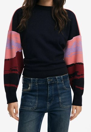Vrouw draagt een zwarte trui met kleurvlakken op de mouwen in roze, rood en paars, gecombineerd met donkerblauwe high-waist spijkerbroek met voorzakken.