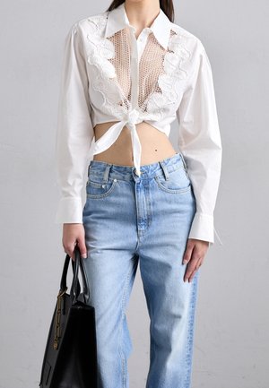 Kvinde iført en hvid blonde- og net-croptop bundet i taljen, lyseblå højtaljede jeans og holder en sort håndtaske mod grå baggrund.