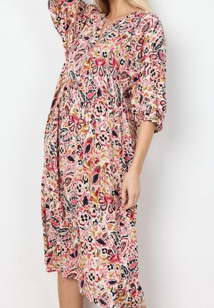 Kobieta w sukience o długości do kolan, luźnym kroju, z różowymi, musztardowymi, czarnymi i białymi kwiatowymi wzorami oraz rękawami o długości 3/4, na białym tle.