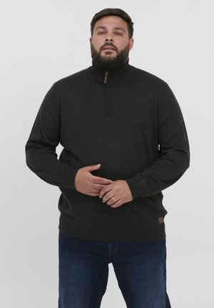 Schwarzer Halbzip-Pullover mit geripptem Kragen und Bündchen, aus weichem Material gefertigt. Mit einem kleinen braunen Leder-Logostick am Saum. Getragen mit blauen Jeans.