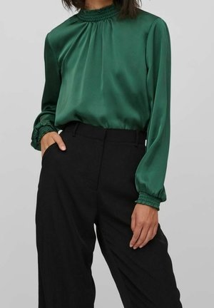 Kobieta ubrana w zieloną satynową bluzkę z długim rękawem, z marszczonym, wysokim dekoltem i mankietami, w połączeniu z czarnymi, szerokimi spodniami z wysokim stanem.
