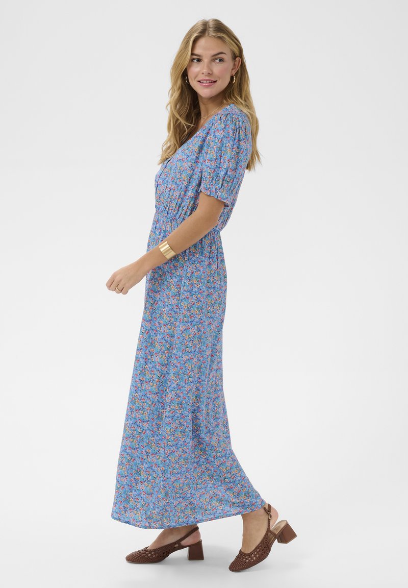 Woman in a blue floral maxi dress wearing brown block-heel shoes and a gold cuff bracelet, looking over her shoulder.