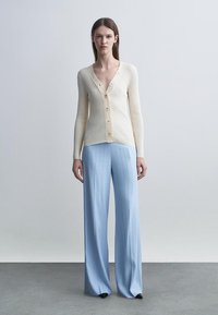 Nainen seisoo päällään kermaisen värinen ribbineulottu napillinen neuletakki ja vaaleansiniset leveälahkeiset housut, taustalla yksivärinen harmaa seinä.