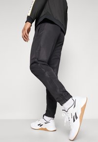 Black athletic joggers with elastic cuffs, featuring side pockets and a subtle logo detail. Paired with white and black athletic shoes.