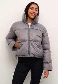 Šedá puffová bunda s vysokým golierom, zapínaním na zips a bočnými vreckami. Má prešívaný dizajn a elastické manžety. Nosená cez biely top.