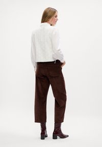 Witte, gestructureerde gilet over een blouse met lange mouwen, gekoppeld aan bruine, corduroy broeken en bordeauxrode enkellaarsjes. Eenvoudig, schoon ontwerp.