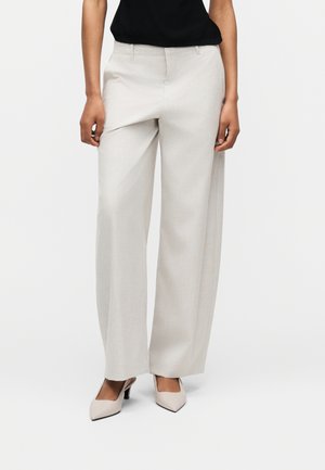 Frau trägt beige weite Hosen, schwarzes Oberteil und beige spitze High Heels, steht vor einem schlichten weißen Hintergrund.