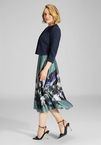 Chaqueta recortada azul con mangas de tres cuartos, combinada con un vestido largo verde con un estampado floral en tonos púrpura, azul y blanco.