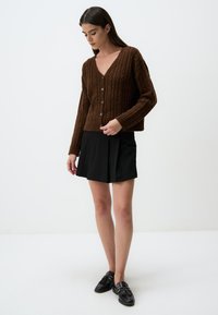 Bruine gebreide cardigan met een V-hals, knoopsluiting en een gestructureerd kabelpatroon, gecombineerd met een zwarte geplooide rok en zwarte loafers.