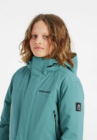 Türkisfarbene Outdoor-Jacke mit hohem Kragen, Reißverschlusstaschen und schwarzen Logo-Akzenten. Hergestellt aus wasserdichtem Material mit glatter Textur.