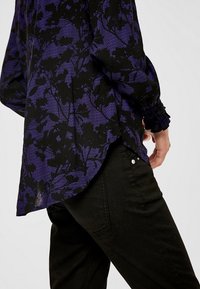 Blusa nera con motivo floreale viola, tessuto leggero, maniche lunghe con polsini elasticizzati. Indossata con pantaloni neri dotati di tasca laterale.