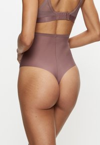 Vrouw met mauve hooggetailleerd naadloos ondergoed en bijpassende beha, gezien van achteren tegen een effen witte achtergrond.