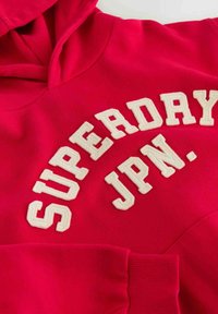 Červená mikina s kapucí a velkým bílým vyšitým logem "SUPERDAY JPN." Obsahuje klokaní kapsu a žebrované manžety. Měkká textura, ležérní design.