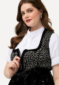 Chaleco de estampado de leopardo negro y gris con cierre de cremallera sobre una blusa blanca plisada, que presenta un cuello redondo y un borde de encaje. Lazo de satén negro en la cintura.