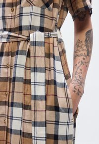 Καφέ και εκρού καρό πουκαμισοφόρεμα με κοντά μανίκια, κλείσιμο με κουμπιά, ζώνη στο ίδιο ύφασμα και πλαϊνές τσέπες. Λεπτομέρειες με ορατή ραφή και υφή.