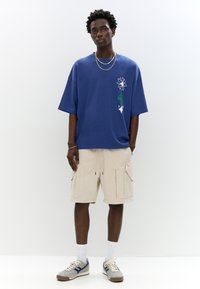 T-shirt blu oversize con stampe grafiche, pantaloni cargo beige con tasche e sneaker grigie con accenti blu. Design minimalista.